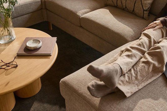 Beige hjørnesofa med puder; person med sokker hviler benene på chaiselongen. Rundt sofabord i træ med vase, briller og bog i en varm, neutral stue.