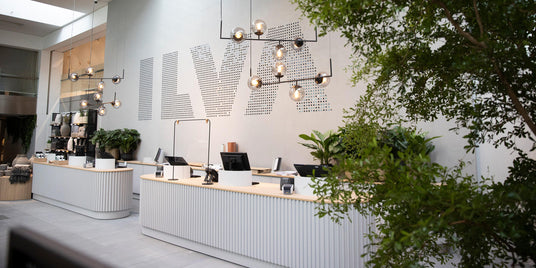 Moderne butikskasse med hængende lamper, grønne planter og stor ILVA‑logo på væggen.