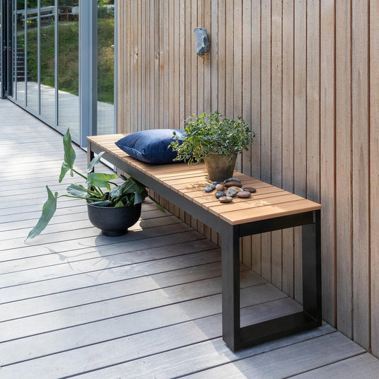 Træterrasse langs en træbeklædt husfacade med bænk i træ og sort metal, blå pude, potteplanter og sten i dagslys.