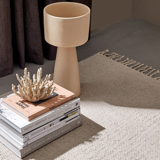 En stak designbøger, bl.a. “THE DESIGN HOTELS BOOK”, “PERFECT IMPERFECT” og “Creative”, toppet af en koralskulptur ved siden af en beige tekstureret bordlampe på et neutralt vævet tæppe med frynser; blødt dagslys og mørke gardiner i baggrunden.