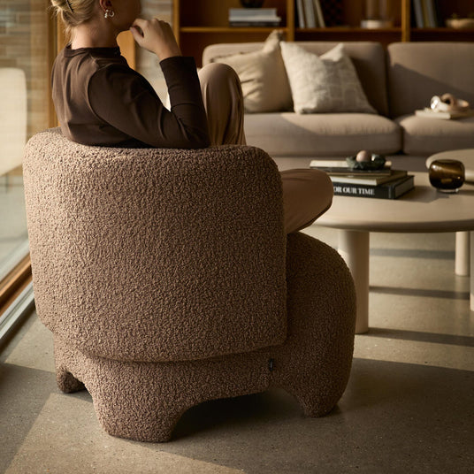 En person sidder i en brun bouclé‑lænestol i en hyggelig stue med beige sofa og rundt sofabord med bøger, hvor en har titlen ”OUR TIME”.