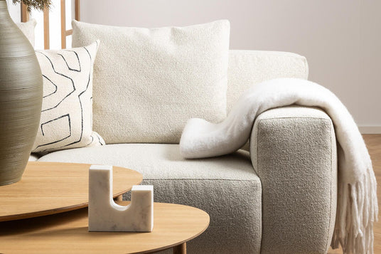 Lys beige bouclésofa med hvid plaid og mønstret pude, foran lyse træborde med marmordekoration og vase i et nordisk stuemiljø.