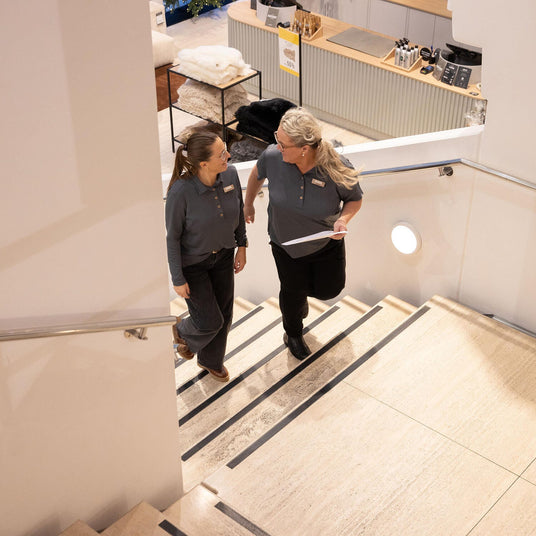 To butiksmedarbejdere går op ad en trappe i en moderne boligbutik; den ene holder et papir; i baggrunden ses et skilt med -50%.