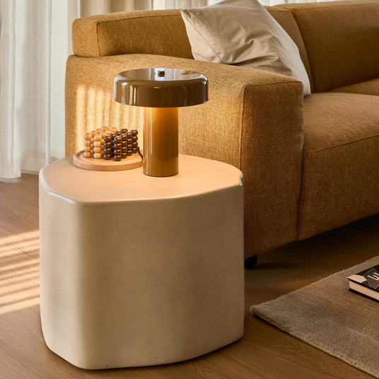 Stue i varme nuancer med skulpturelt beige sidebord, tændt karamelfarvet svampelampe, træperler på bakke og sennepsgul sofa med pude ved lette gardiner i dagslys.
