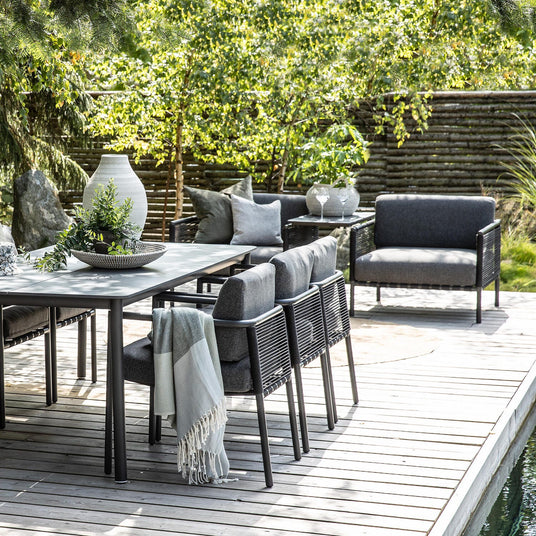 Udendørs møbelsæt på træterrasse: sort spisebord med grå polstrede stole, loungestol og sofa; bordet stylet med hvid vase, skål med grønne grene og en lys plaid, omgivet af frodig have.