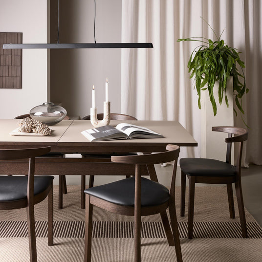 Minimalistisk spisestue med mørke træstole, beige bord, pendellampe, tændte lys, glasvase, åben bog og høj grøn plante foran lyse gardiner.