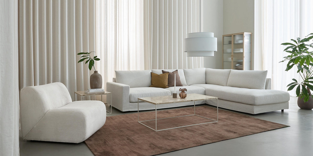 Lys minimalistisk stue med hvid hjørnesofa og loungestol, brunt tæppe, slankt stenbord, stor hvid pendellampe, høje beige gardiner og grønne potteplanter.