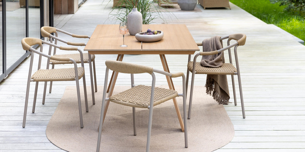 Udendørs spiseområde på træterrasse: firkantet træbord med fletstole, beige udemåtte, vase med blomster, skål med frugt og vinglas; loungemøbler og grøn have i baggrunden.