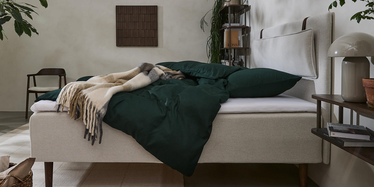 Lyst soveværelse med beige polstret seng, mørkegrønt sengetøj, uldtæppe og planter.