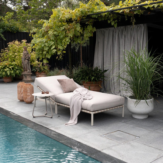 Poolsiden med beige liggestol med pude og strikket plaid, lille sidebord og rattanlanterner under en pergola med vinranker og krukplanter ved en pool.