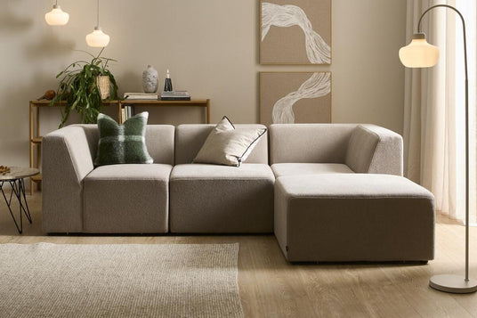 Beige modulsofa i lys skandinavisk stue med gulvlampe, tre pendler, vægkunst, tæppe og grøn plante.