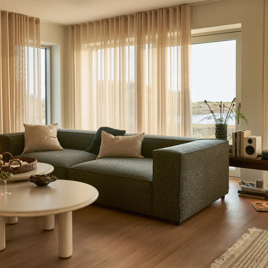Lyst stue med transparente beige gardiner, mørkegrå modulsofa med puder, runde hvide sofaborde og planter ved store vinduer med udsigt til vand.