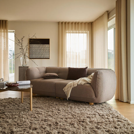 Lyst stue med beige, transparente gardiner, buet taupe-sofa, langhåret tæppe og træsofabord med bog; plaid og puder skaber en varm, skandinavisk stemning.