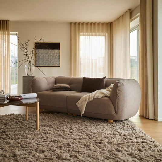 Lyst stue med beige, transparente gardiner, buet taupe-sofa, langhåret tæppe og træsofabord med bog; plaid og puder skaber en varm, skandinavisk stemning.