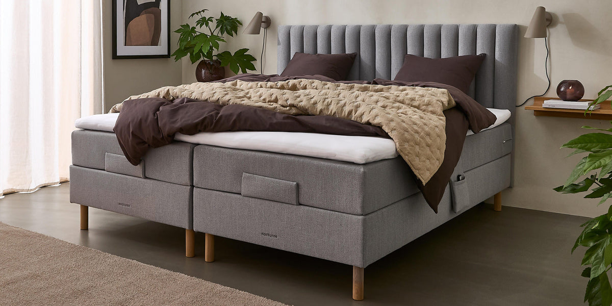 Lyst soveværelse med grå kontinentalseng, beige quiltet plaid og mørkebrunt sengetøj, væglamper, plante og vægkunst ved et stort vindue med lyse gardiner.