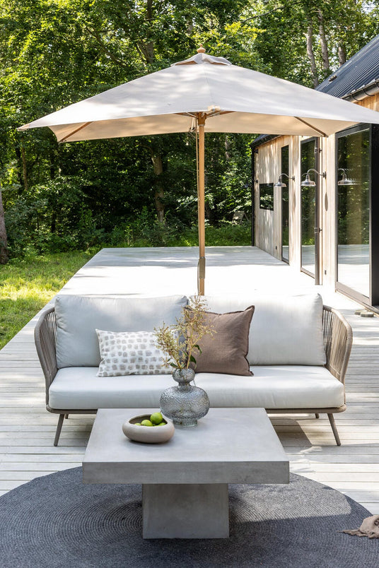 Lys træterrasse med beige parasol, sofa med puder og betonbord med vase og skål med lime foran et moderne træhus ved skovkanten.