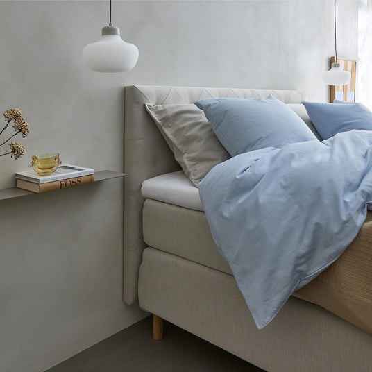 Lyst soveværelse med beige seng, lyseblåt sengetøj og puder, to hvide pendler samt en smal hylde med vase, gul kop og bogen FOSS ved siden af sengen.