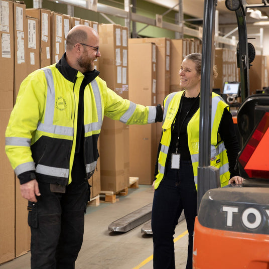 To lagermedarbejdere i gule refleksveste ved en orange truck med ”TOYO” logo, omgivet af stablede papkasser i et indendørs lager.