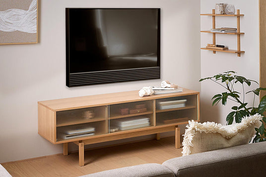 Lyst skandinavisk stue med vægmonteret TV over et lavt mediemøbel i lyst egetræ, grå sofa, lille hylde og grøn potteplante i dagslys.