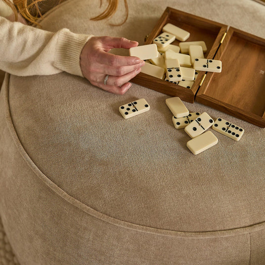 Hånd tager en dominobrik fra en trææske på en beige puff/ottoman.