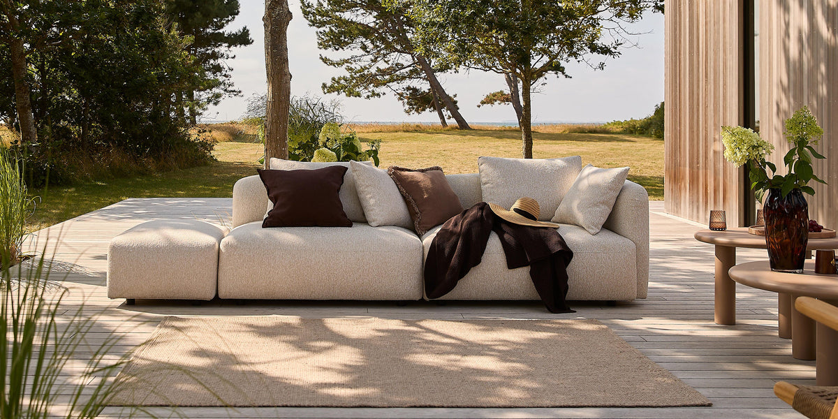 Lys terrasse med beige modulsofa med brune puder, plaid og stråhat, neutral tæppe og sideborde med vase og grønne planter foran et træbeklædt hus i dagslys.