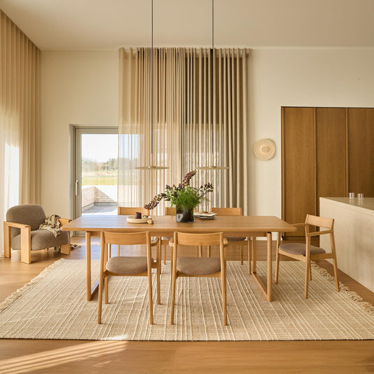 Lys og rolig spisestue i skandinavisk/Japandi-stil med egetræsbord og fire stole på lys vævet tæppe, to pendler over, beige gardiner og en vase med grønt som midtpunkt; loungestol ved siden af.