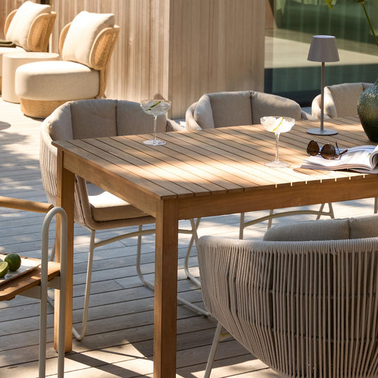 Solbeskinnet terrasse med teakbord og polstrede rebstole; to cocktailglas med citrus, en lille bærbar bordlampe, solbriller og et opslået magasin på bordet.