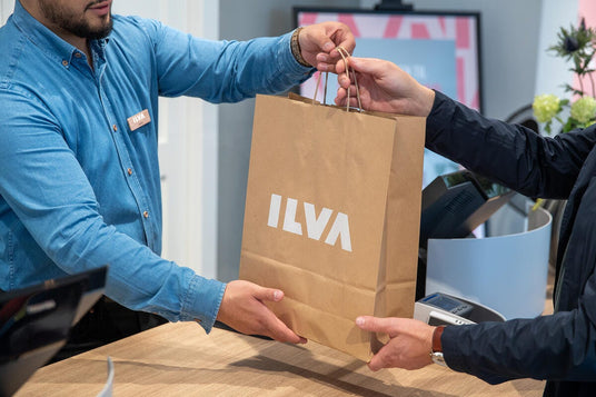 Kunde og medarbejder rækker hinanden en brun papirpose med ILVA‑logo ved kassen.