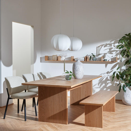 Lyst skandinavisk spisestue med rillet træbord og bænk, lyse stole, to hvide pendler, hylde med dekorationer samt grønne planter og lysestager.