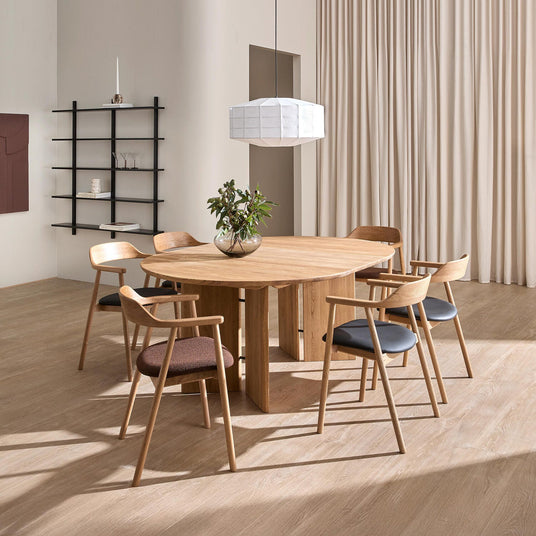 Minimalistisk spisestue med rundt egetræsbord, seks træstole, hvid pendel og glasvase med grønne grene; beige gardiner og sort hylde i baggrunden.
