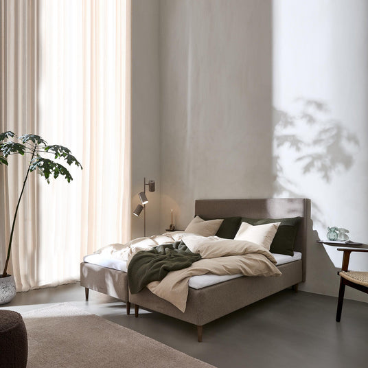 Lyst soveværelse med polstret seng i beige og grønne nuancer, lette gardiner, væglampe, sidebord med glasvase og en stor plante i hjørnet.