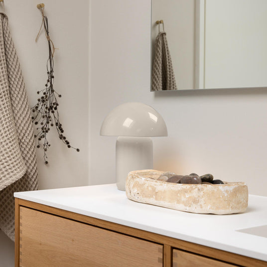 Badeværelse med træskab og hvid bordplade, svampeformet bordlampe og stentruge med små sten ved siden af spejl og ophængt håndklæde.