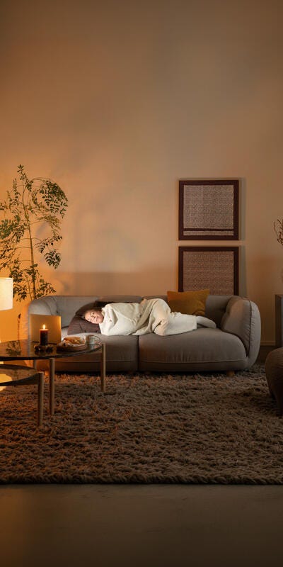 Stue i varm belysning med en person, der sover på en grå sofa under et tæppe; sofaborde med tændt lys, kop og snack, gulvlampe, vægkunst, stor blød måtte og planter.