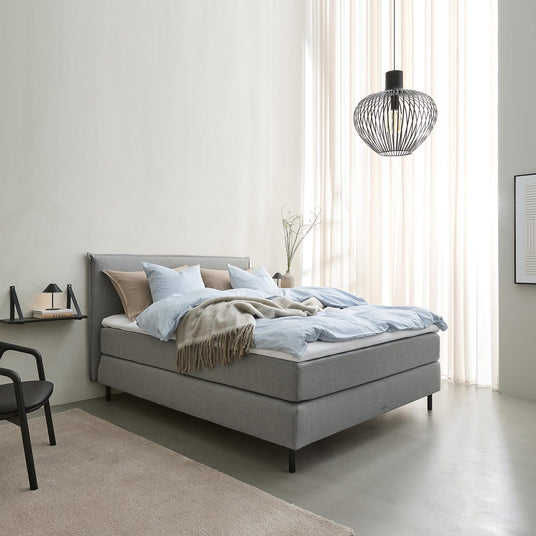 Lyst soveværelse i skandinavisk stil med grå polstret seng, lyseblåt sengetøj og beige plaid, sort trådpendel og lette gardiner, der slipper dagslys ind.