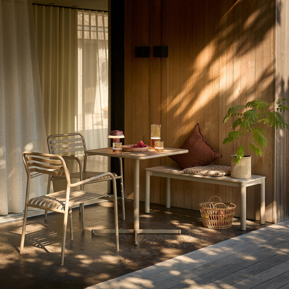Lille terrasse med beige metalstole, kvadratisk bord og bænk ved en trævæg; på bordet står en karaffel og glas med drikke, en lille bærbar lampe og en skål med bær; plettet sollys falder over scenen.