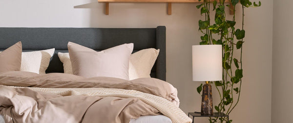 Hyggeligt soveværelse med grå polstret seng, beige sengetøj og plaid, sengebord med lampe og bøger, hylde med dekor og hængeplante samt grøn puf.