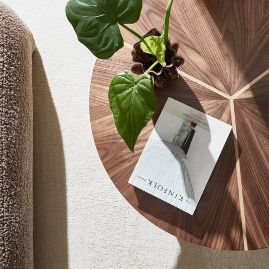 Rundt sofabord i valnød med stjernefinér, en grøn potteplante og bogen THE KINFOLK HOME på bordpladen i blødt dagslys.