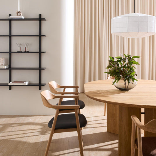 Lyst spisestue i minimalistisk stil med rundt egetræsbord, træstole med sorte sæder, hvid pendellampe, beige gardiner og en grøn plante i glasvase på bordet; til venstre en sort væghylde med bøger, glas og en slank lysestage.