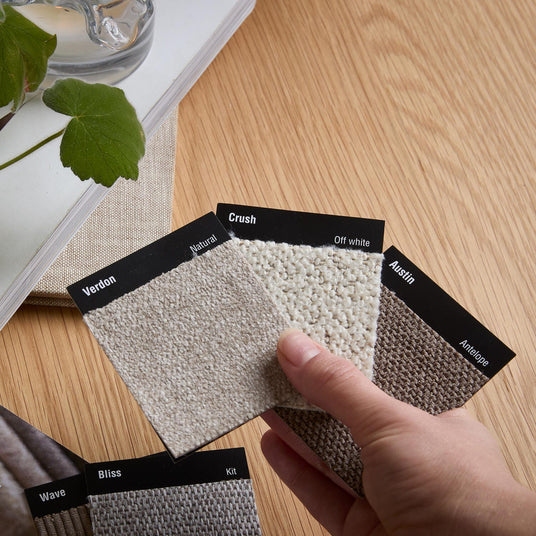 En hånd holder stofprøver med teksten: Verdon – Natural, Crush – Off white, Austin – Antelope; flere prøver ved siden af mærket Wave, Bliss og Kit på et træbord; et magasin ved siden af siger: ”Et lille stofprøve i hånden kan være begyndelsen på noget stort”.