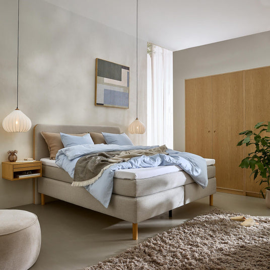 Roligt skandinavisk soveværelse med lysegrå seng, lyseblåt sengetøj og beige plaid, to hængende lamper, svævende sengebord, vægkunst, garderobe i træ, shaggy tæppe og grøn plante i blødt dagslys.