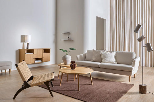 Lys, minimalistisk stue med beige sofa, træ-sofaborde, brun tæppe, fletlænestol og standerlampe foran høje gardiner.
