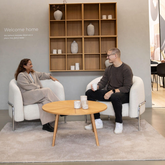 Moderne showroom med to personer i hvide lænestole omkring et rundt sofabord på et lyst tæppe, væghylde med vaser bagved og budskabet ”Welcome home – We believe everyone deserves a place they feel at home”, en tredje person går forbi i baggrunden.