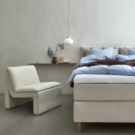 Lyst, minimalistisk soveværelse med beige polstret seng med lyseblåt sengetøj og brun plaid, en cremefarvet loungestol, hængende hvid pendellampe og smal væghylde med vase, gren og tændt lys.