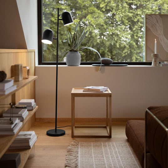 Minimalistisk stuekrog med sort gulvlampe med to skærme, åbne træhylder med bøger, et lille kubeformet sidebord i eg og et lyst tæppe; i vindueskarmen står en potteplante, et lys og en ramme, med dagslys og grøn udsigt udenfor.