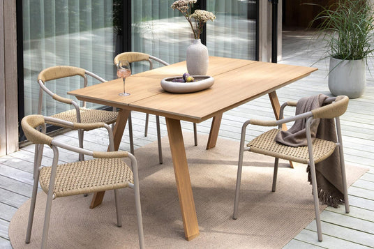 Terrasse med spisebord i træ og fire fletstole på trægulv, vase med blomster og skål på bordet, plaid over en stol og krukkeplante i baggrunden i dagslys.