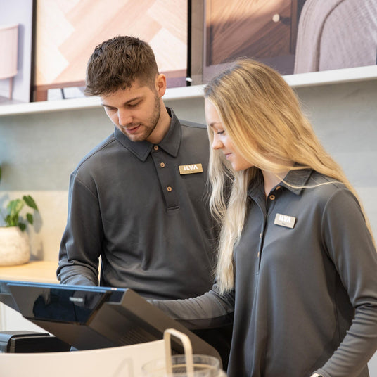 To butiksmedarbejdere i gråt ILVA-tøj ved kassen, kigger på en POS-skærm i et moderne showroom med grønne planter i baggrunden.