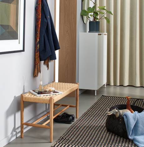 Entré med fletbænk i træ, ophængt frakke og tørklæde, bakke med duftpinde, hvidt skab med potteplante, beige gardiner og mønstret tæppe; kurv med legetøj og blå plaid.