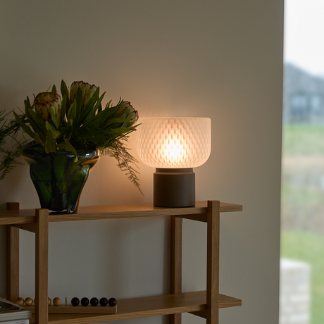 Tændt bordlampe i frostet glas på en træreol ved siden af en grøn vase med blomster ved et vindue.