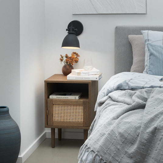 Soveværelse med grå seng, blå- og gråt sengetøj og plaid, et natbord i træ med rottinglåge, lille vase med tørrede blomster, sort væglampe og en stak bøger – Kinfolk Travel, Simplicity at Home og Be Toogood – samt et glas vand ovenpå.