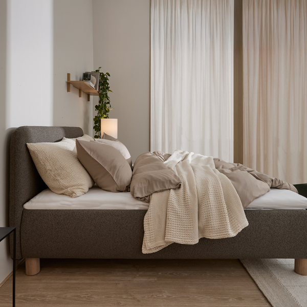 Lyst soveværelse med polstret grå seng, beige sengetøj og cremefarvet plaid, lette gardiner foran vinduerne samt en lille bordlampe og hængende grøn plante.